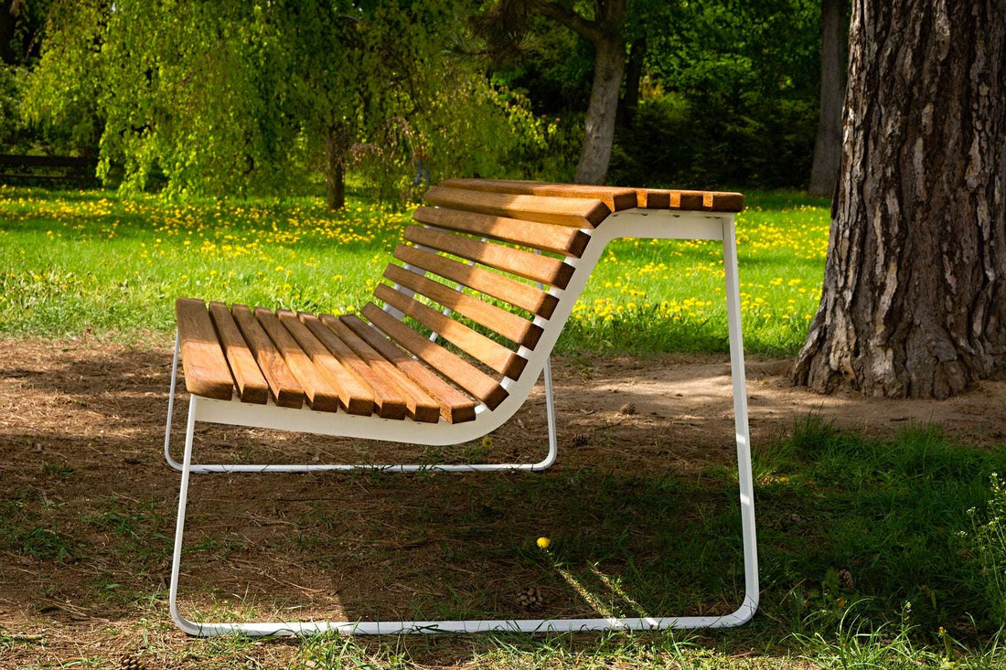 Moderne Parkbank aus Holzlatten mit weißem Metallrahmen, aufgestellt in einem grünen Park mit Bäumen und Blumen im Hintergrund.