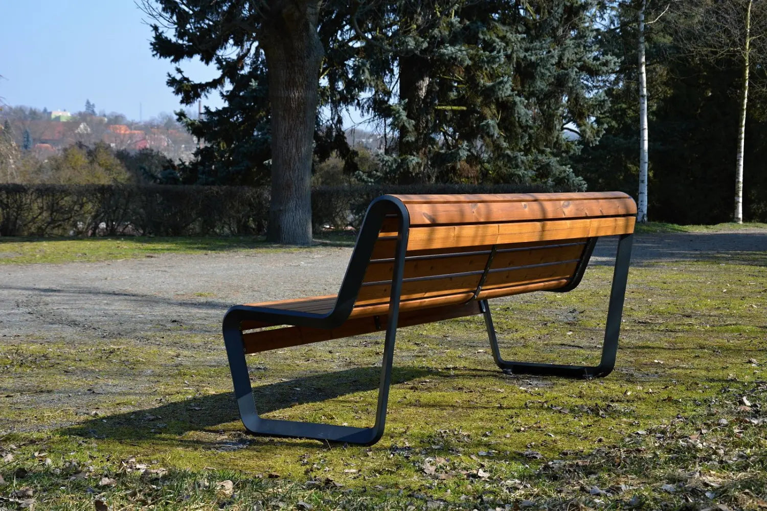 Eine moderne Parkbank aus Holz mit schwarzem Metallgestell steht auf einer Wiese, im Hintergrund sind Bäume und Sträucher.