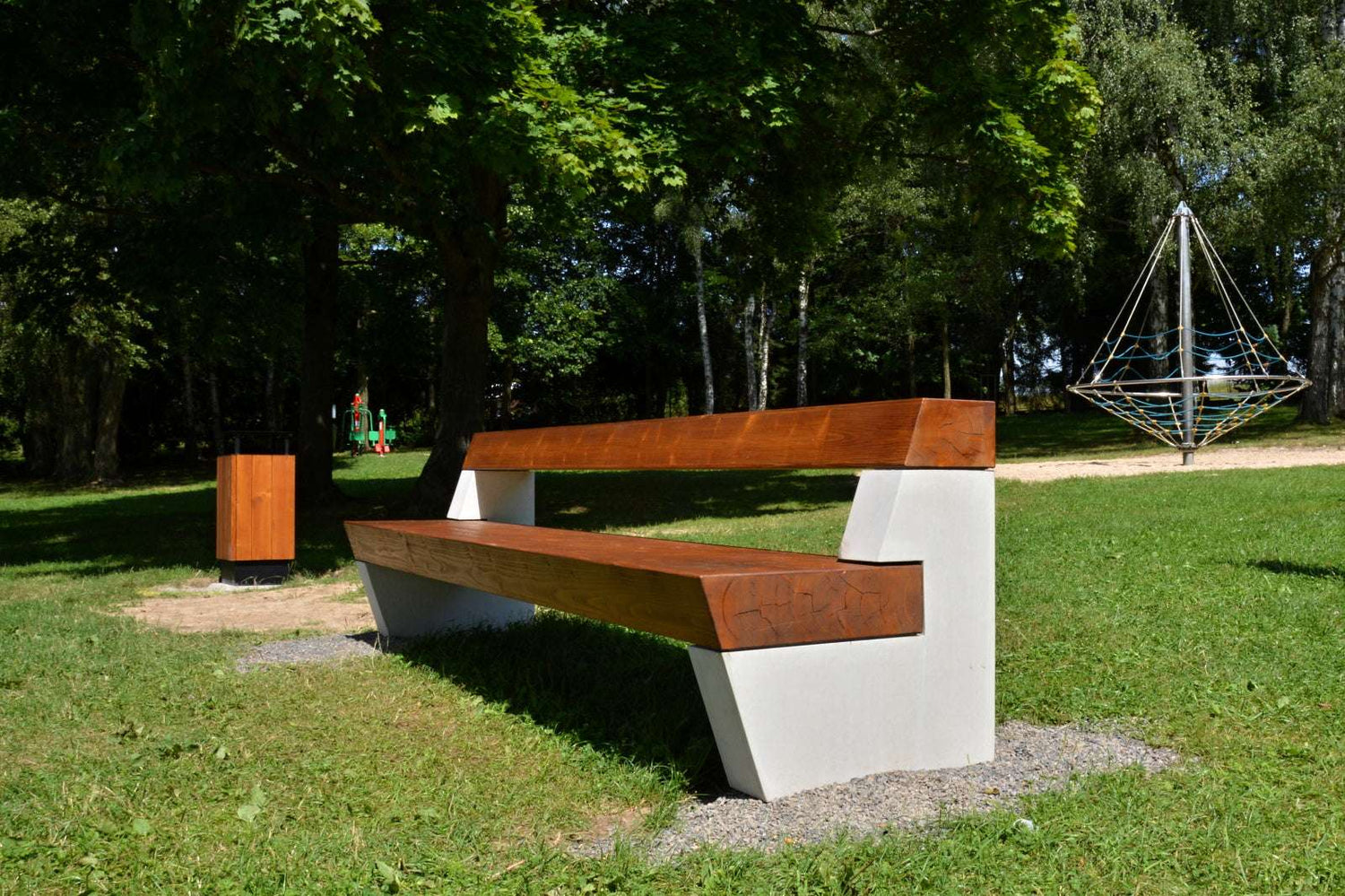 Moderne Parkbank aus Holz und Beton auf grüner Wiese im sonnigen Park mit Bäumen und Spielplatz im Hintergrund.
