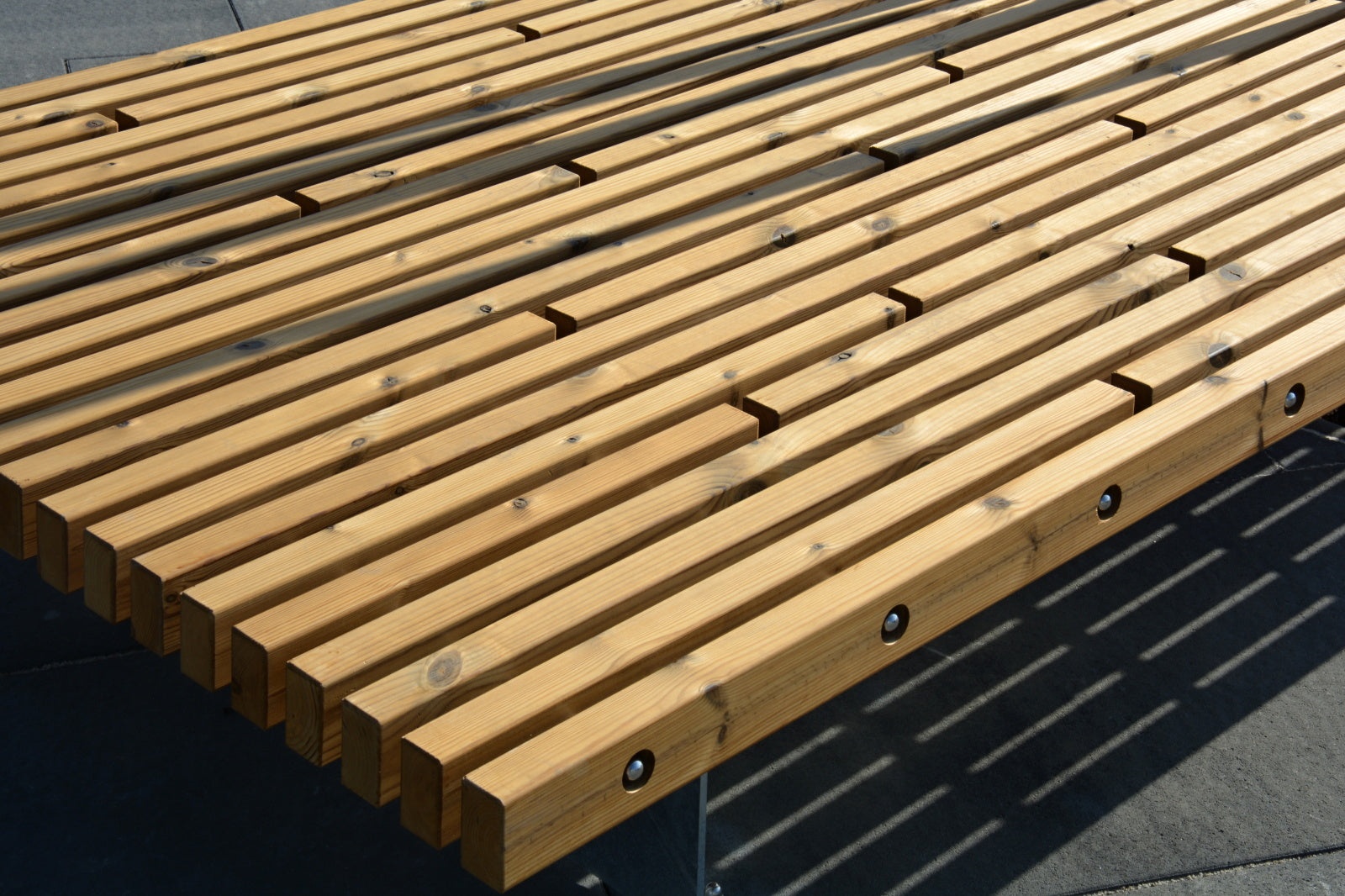 Holzbank im modernen Design mit parallel verlaufenden Latten und Metallbefestigungen auf einem gepflasterten Untergrund.