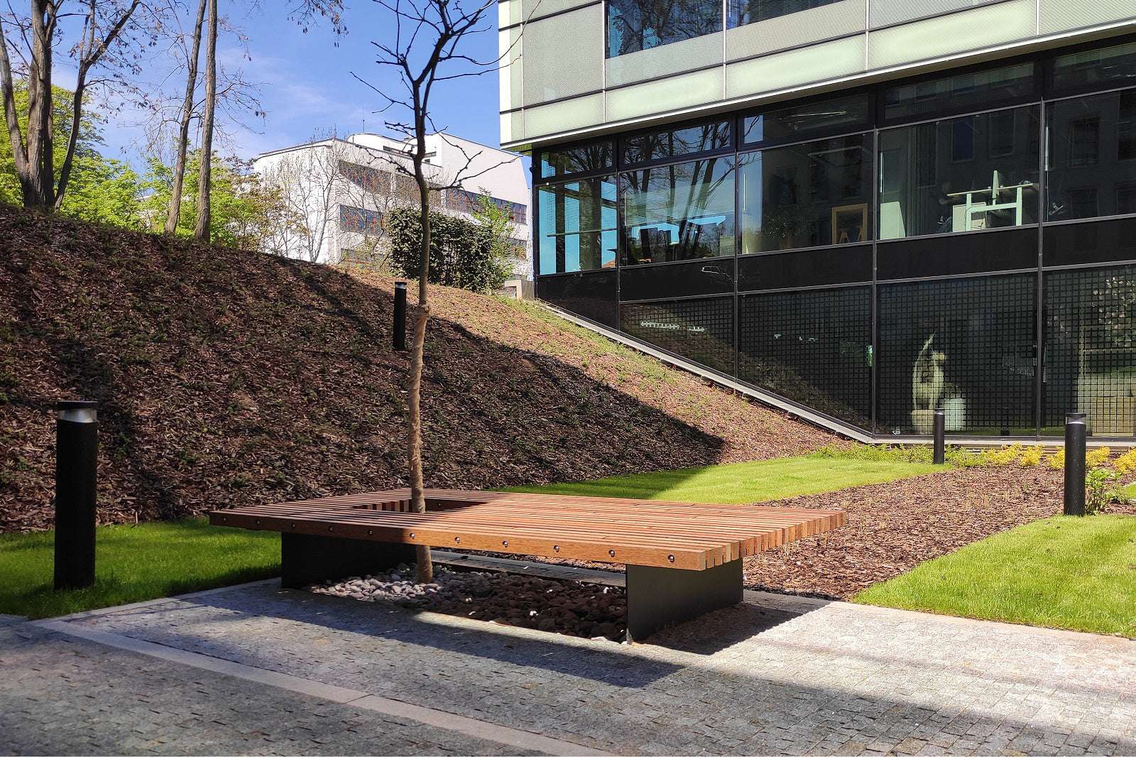 Moderne Holzbank vor einem Bürogebäude, platziert auf einem Pflasterweg mit begrünter Umgebung und kleiner Baum.