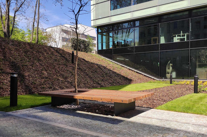 Moderne Holzbank vor einem Bürogebäude, platziert auf einem Pflasterweg mit begrünter Umgebung und kleiner Baum.