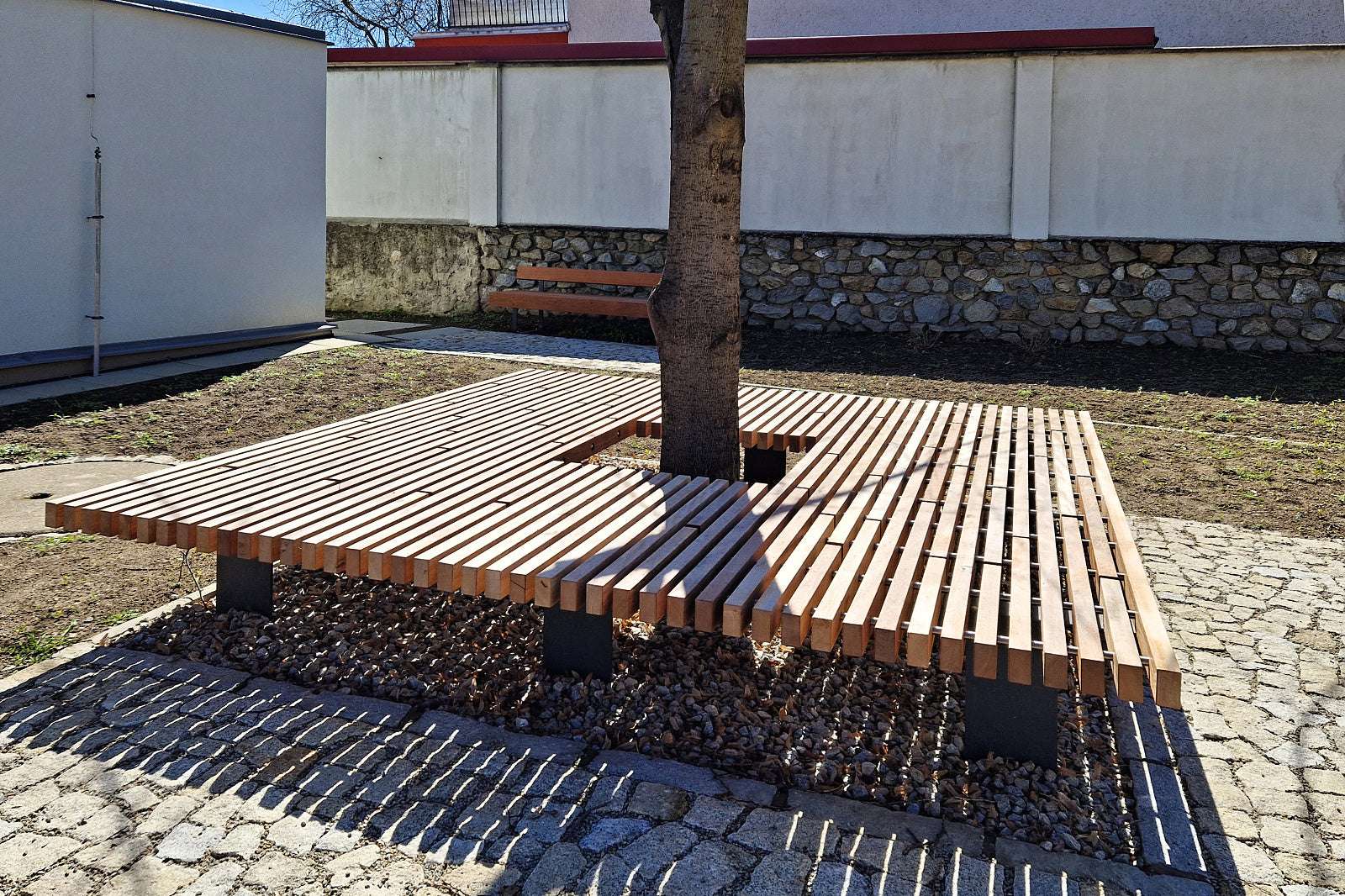 Holzbank um einen Baum herum auf gepflastertem Boden in einem urbanen Umfeld.