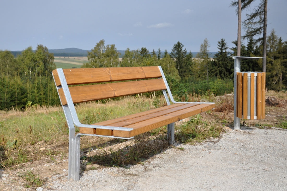 Holzbank mit Metallrahmen neben einem Mülleimer in einer ländlichen Landschaft mit Bäumen und Hügeln im Hintergrund.