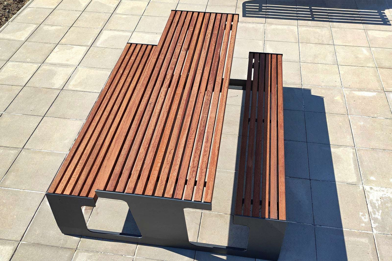 Moderne Gartenbank aus Holz und Metall auf gepflastertem Boden bei Sonnenschein.