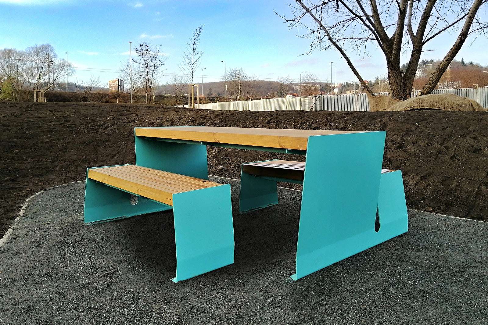 Moderne blaue und holzfarbene Picknickbank auf einem Kiesboden, umgeben von kahlen Bäumen und Baustelle.