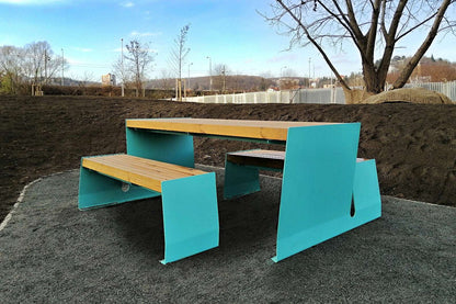 Moderne blaue und holzfarbene Picknickbank auf einem Kiesboden, umgeben von kahlen Bäumen und Baustelle.
