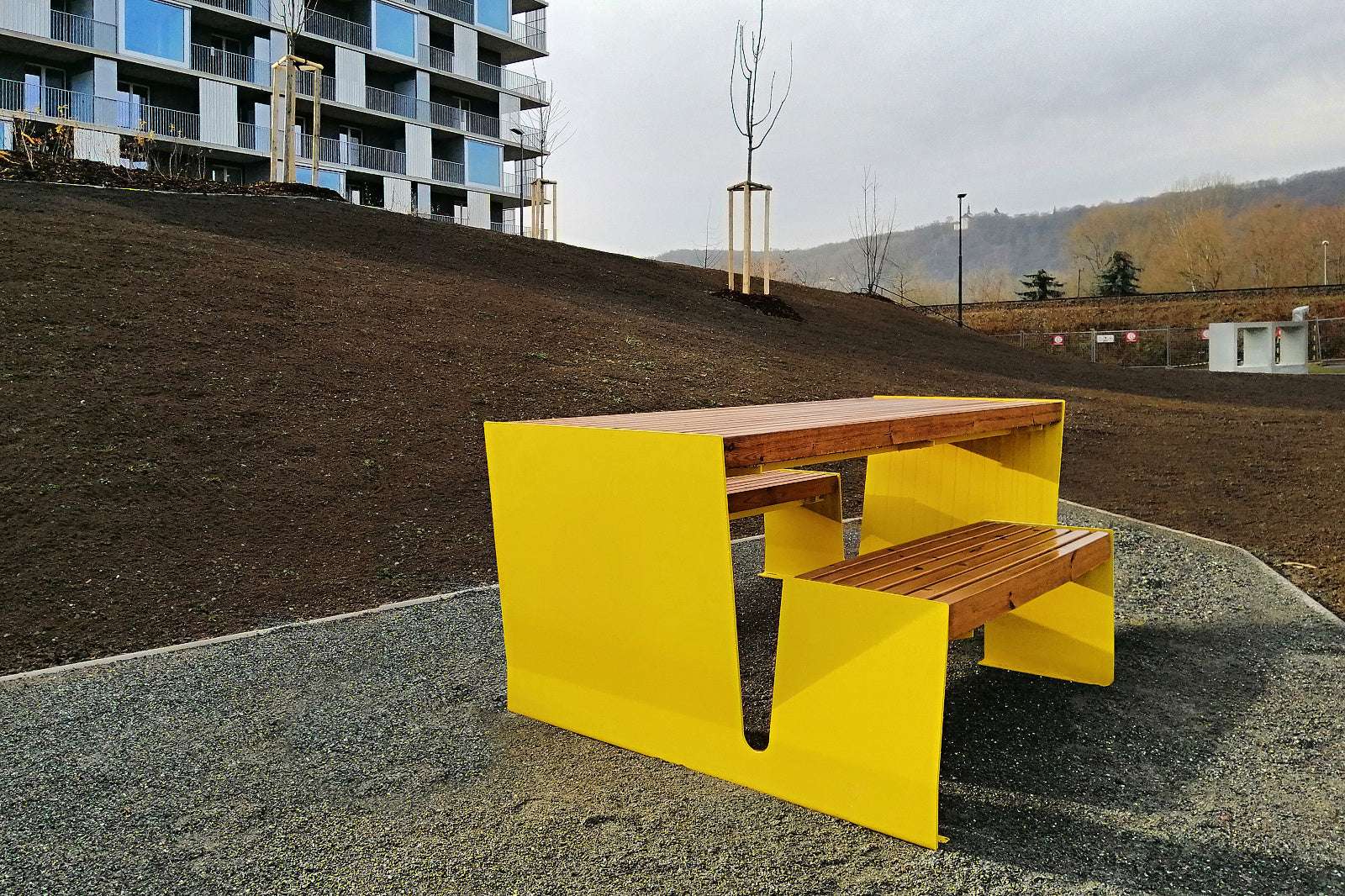 Moderne gelbe Parkbank mit Tisch auf Kiesweg vor Neubau und begrüntem Hügel.