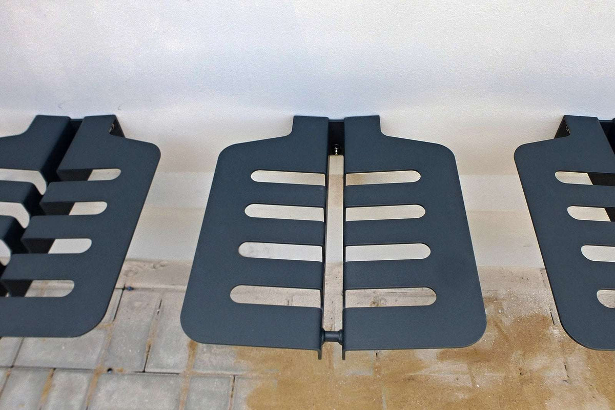 Auf dem Bild sind drei nebeneinander angeordnete moderne Sitzflächen aus dunklem Metall zu sehen. Die Sitzflächen haben breite, schlitzenförmige Aussparungen und sind an einer Wand befestigt. Der Boden darunter ist teilweise gepflastert und teilweise mit Sand oder Erde bedeckt. Die Struktur wirkt minimalistisch und funktional, möglicherweise als Sitzgelegenheit für den Außenbereich konzipiert.