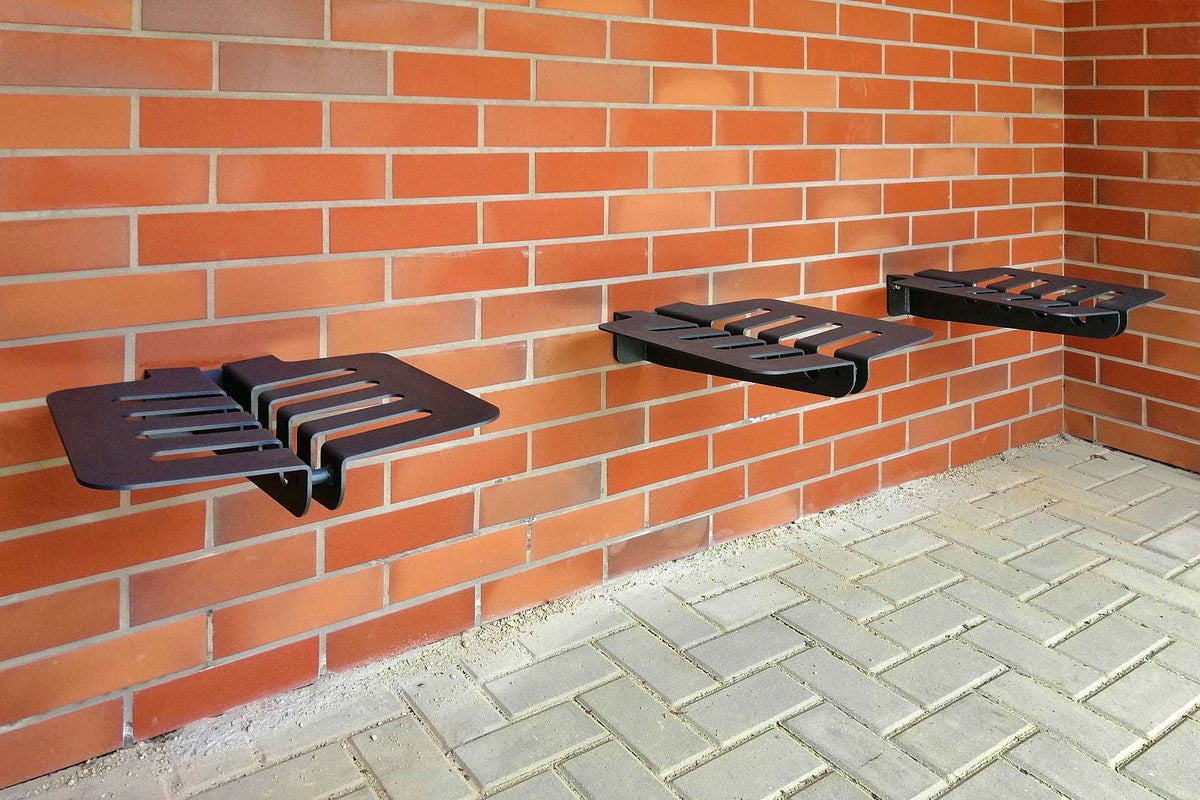 Zu sehen sind drei schwarze Metallbänke mit modernen, rechteckigen Aussparungen, die an einer roten Ziegelmauer montiert sind. Der Boden besteht aus hellgrauen Pflastersteinen. Die Bänke scheinen fest an der Wand verschraubt zu sein und schweben leicht über dem Boden. Die gesamte Szenerie wirkt ordentlich und ist wahrscheinlich Teil eines Außenbereichs, etwa vor einem Gebäude oder in einem Hof.