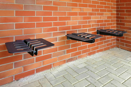 Zu sehen sind drei schwarze Metallbänke mit modernen, rechteckigen Aussparungen, die an einer roten Ziegelmauer montiert sind. Der Boden besteht aus hellgrauen Pflastersteinen. Die Bänke scheinen fest an der Wand verschraubt zu sein und schweben leicht über dem Boden. Die gesamte Szenerie wirkt ordentlich und ist wahrscheinlich Teil eines Außenbereichs, etwa vor einem Gebäude oder in einem Hof.