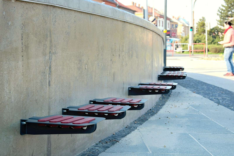 Mehrere Klappsitze aus Metall und Holz mit roten Sitzflächen sind an einer gebogenen Betonwand montiert. Im Hintergrund sieht man einen Gehweg, einen Mann mit roter Jacke und Jeans sowie einige Gebäude, Bäume und Verkehrsschilder. Die Sitze sind in Reihe parallel zur Wand angebracht.