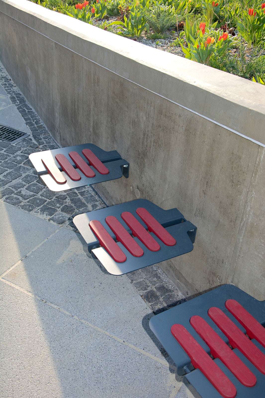 Auf dem Bild sind drei moderne Sitzflächen aus Metall mit roten Polstern an einer Betonmauer montiert. Im Hintergrund blühen rote Tulpen und grüne Pflanzen in einem Beet. Der Boden besteht aus Kopfsteinpflaster und großen Betonplatten.