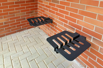 Das Bild zeigt zwei schwarze, modern gestaltete Metallsitze, die an einer orangenen Ziegelwand montiert sind. Die Sitze haben mehrere Schlitze und abgerundete Ecken. Der Boden besteht aus grauen, rechteckigen Pflastersteinen im Fischgrätmuster. Die Umgebung wirkt sauber und minimalistisch, möglicherweise handelt es sich um einen Wartebereich im Freien oder einen öffentlichen Aufenthaltsbereich.