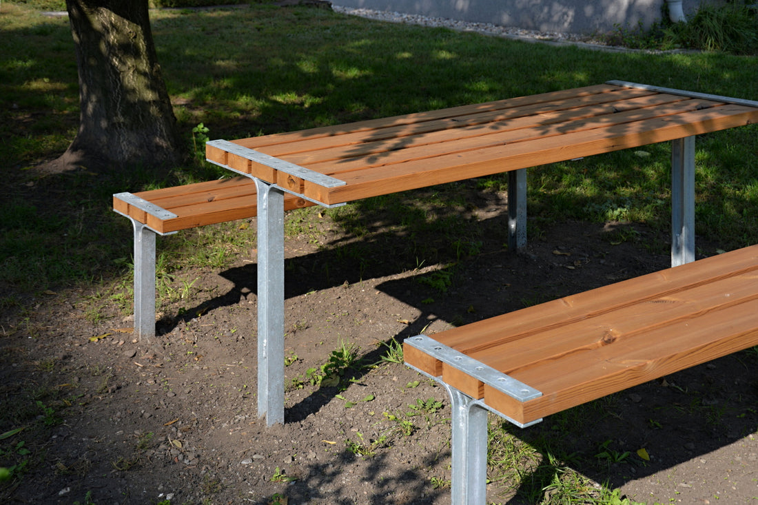 Holztisch mit zwei Bänken auf Metallgestellen im Freien, teilweise im Schatten eines Baumes auf grasbewachsenem Untergrund.