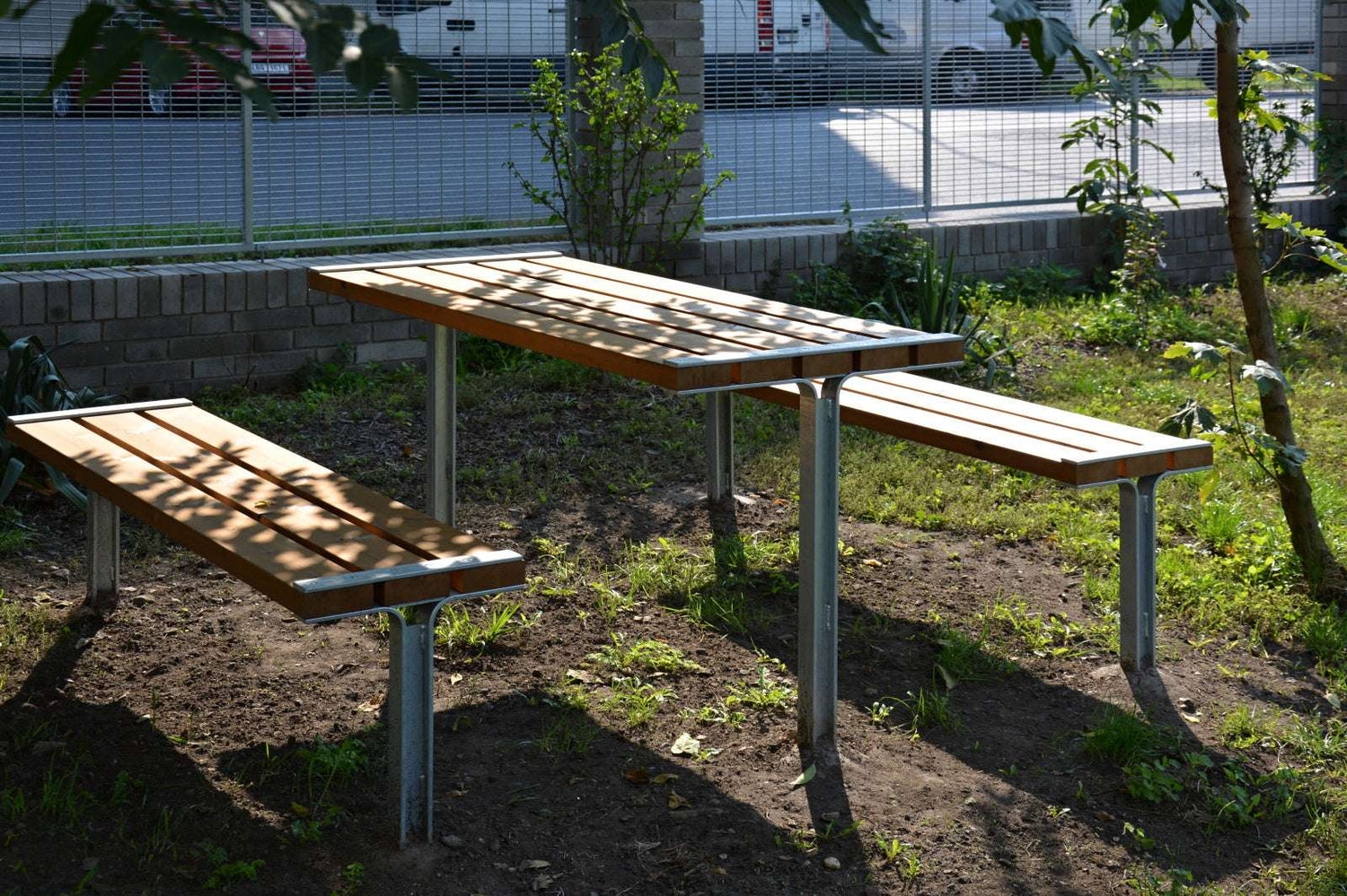 Garten-Tisch-und-Bank-Set aus Holz und Metall auf Grasfläche vor einem Zaun.