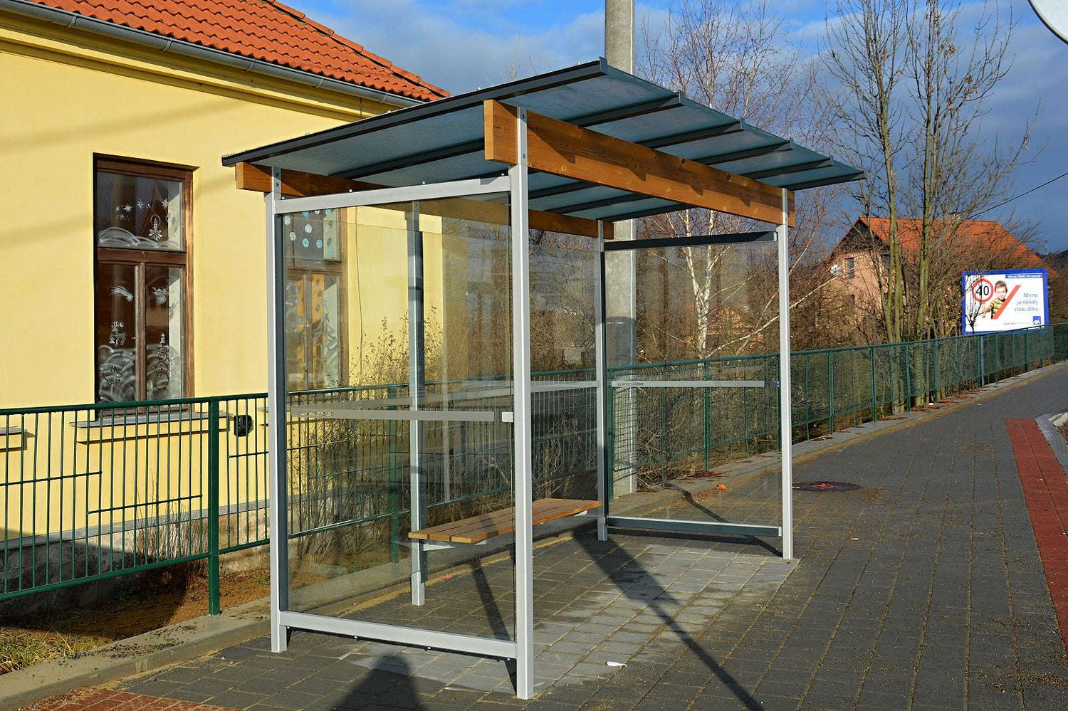 Bushaltestelle mit Glaswänden und Holzdach neben gelbem Gebäude.