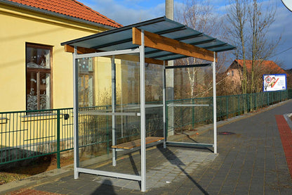 Bushaltestelle mit Glaswänden und Holzdach neben gelbem Gebäude.