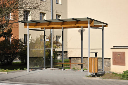 Eine moderne Bushaltestelle mit Glaswänden, einem Holzdach und einer Holzsitzbank, umgeben von Häusern und Grünflächen.
