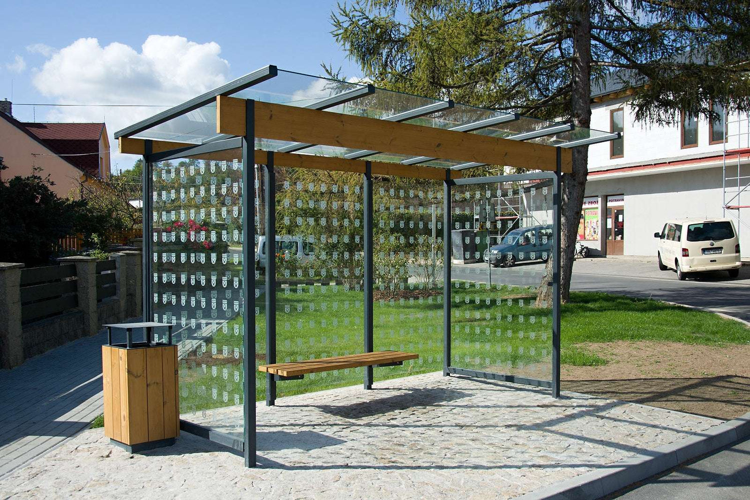 Moderne Wartehalle mit Glasdach und Sitzbank, umgeben von Bäumen und Gebäuden im Hintergrund.