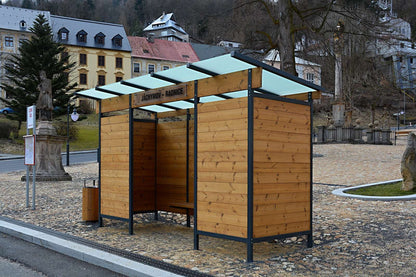 Eine moderne Wartehalle aus Holz und Metall auf einem gepflasterten Platz mit umliegenden Gebäuden.